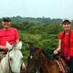 Two people wearing red shirts and hats are riding horses outdoors, surrounded by lush green vegetation and hills—the perfect scene for travel planning as you explore new destinations.