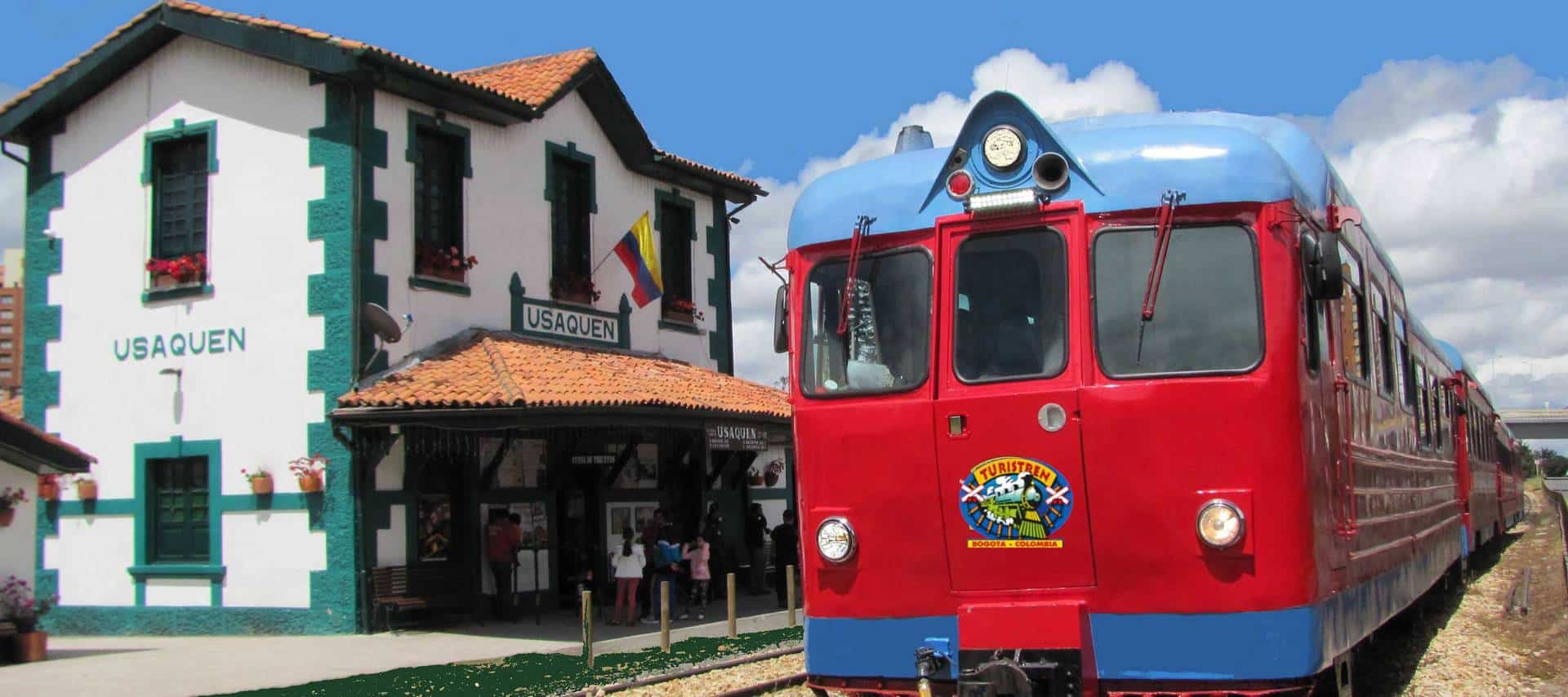 A bright red and blue vintage train is stopped at a station with a white building labeled Usaquen in Colombia. People gather near the entrance under a blue sky with scattered clouds, ready to enjoy scenic train tours.