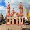 A large, ornate church with twin red-and-white towers stands in a city square in Colombia, surrounded by people and modern buildings under a partly cloudy sky—a perfect sight for a last minute trip.