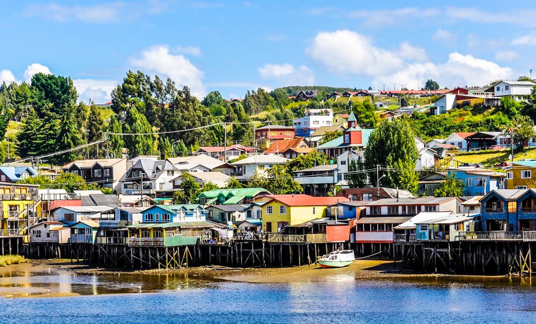 Colorful wooden stilt houses line the waterfront in a small coastal town, with green trees and hillside homes behind, under a bright blue sky—an inviting scene near Refúgia Chiloe Spa and the charming Chiloe Hotel.