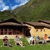 Five people relax on deck chairs on a grassy lawn in front of rustic, thatched-roof buildings, their sense of cooperation reflected in the peaceful setting beneath green mountains and a partly cloudy sky.