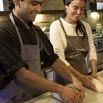 Two people in aprons smile as they knead dough together on a kitchen counter, suggesting they are baking or cooking in a warmly lit kitchen.