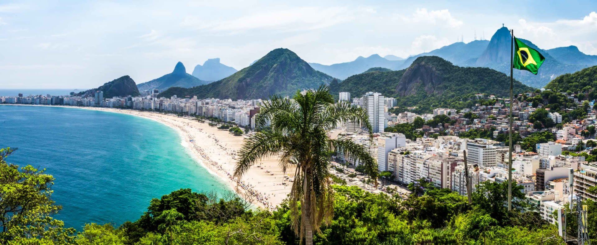 Panoramic view of Copacabana Beach in Rio de Janeiro, Brazil, with city buildings, lush hills, and a Brazilian flag waving in the sun—a must-see for travelers exploring Brazil visa requirements.