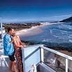 A couple enjoys a wonderful time on a balcony overlooking a sandy beach and ocean waves, with mountains in the background and clear blue skies above.