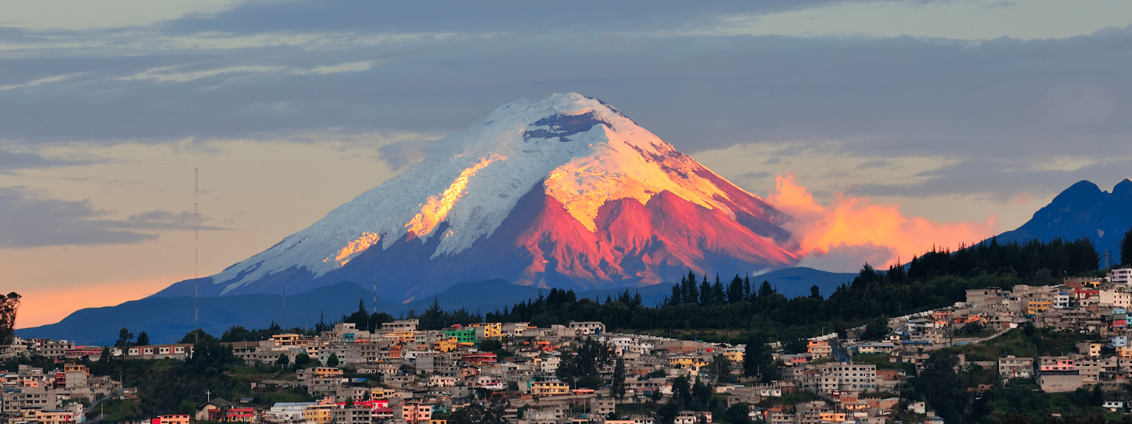 Snow-capped Cotopaxi volcano in Ecuador glows with orange sunlight at dusk, rising above a town with clustered houses and green trees under a partly cloudy sky—an essential scene for any travel guide.