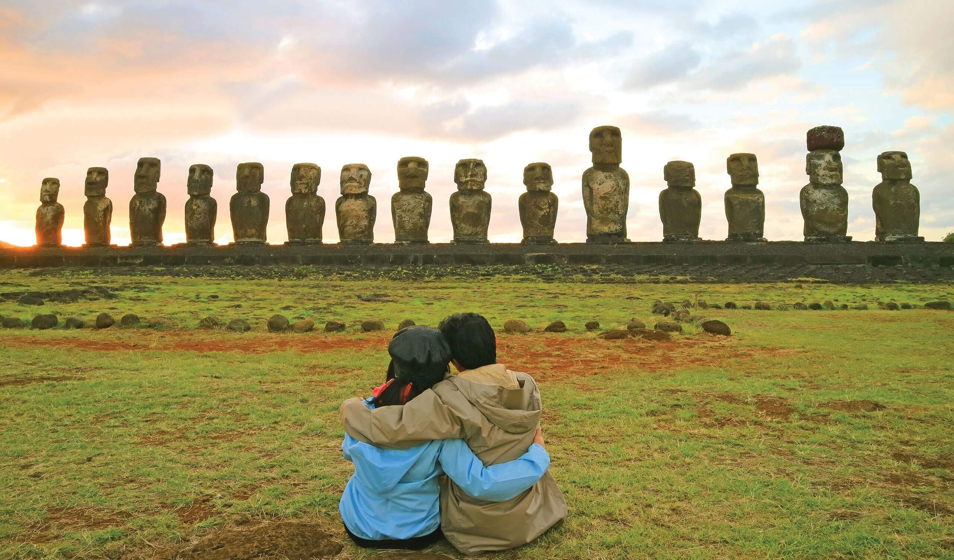 Two people in jackets sit on grass, embracing as they face a row of majestic moai statues at sunrise or sunset on Easter Island—a breathtaking moment that captures the magic of Chile tourism.