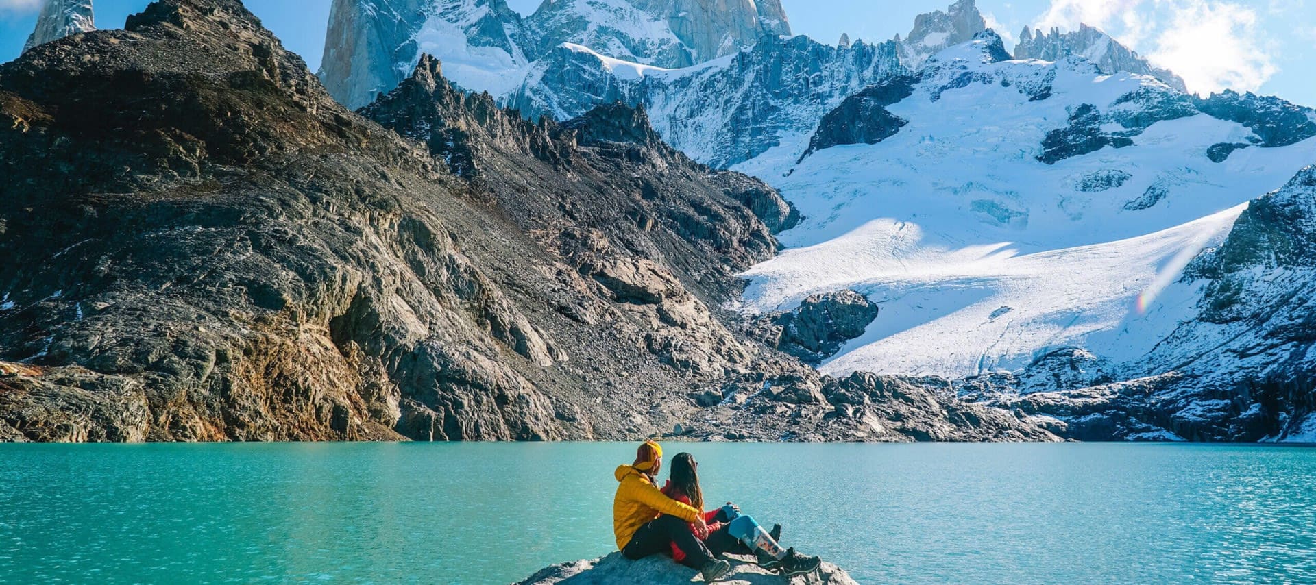Two people in outdoor gear sit on a rock by a turquoise alpine lake in Patagonia, surrounded by rugged mountains and snow-covered peaks under a blue sky—a perfect spot for adventure tours or a memorable honeymoon.