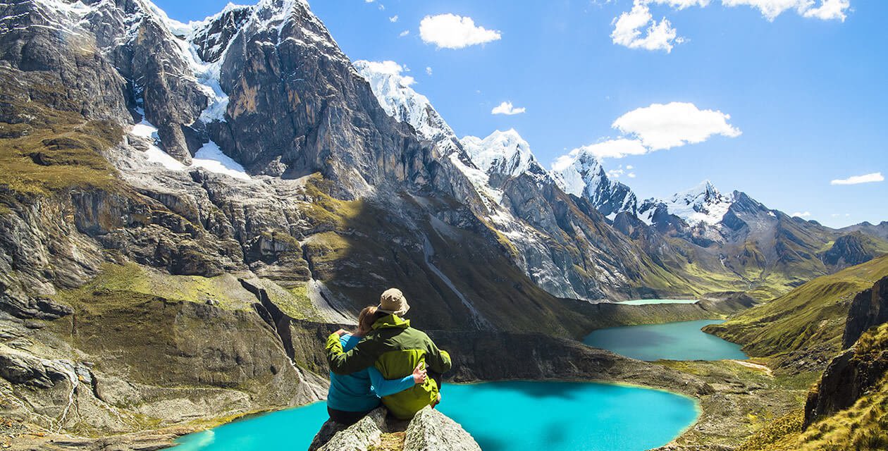 Two people sit on a rocky ledge overlooking turquoise lakes and snow-capped mountains under a bright blue sky, savoring the scenic, rugged landscape—an unforgettable moment perfect for Peru honeymoon memories.