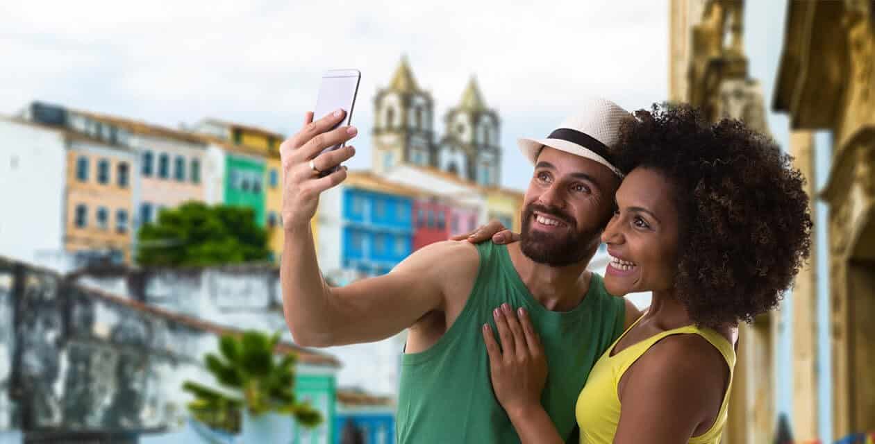A smiling couple takes a selfie together outdoors, enjoying their Brazil honeymoon with colorful historic buildings and a church in the background—a perfect romantic getaway in this vibrant, scenic location.