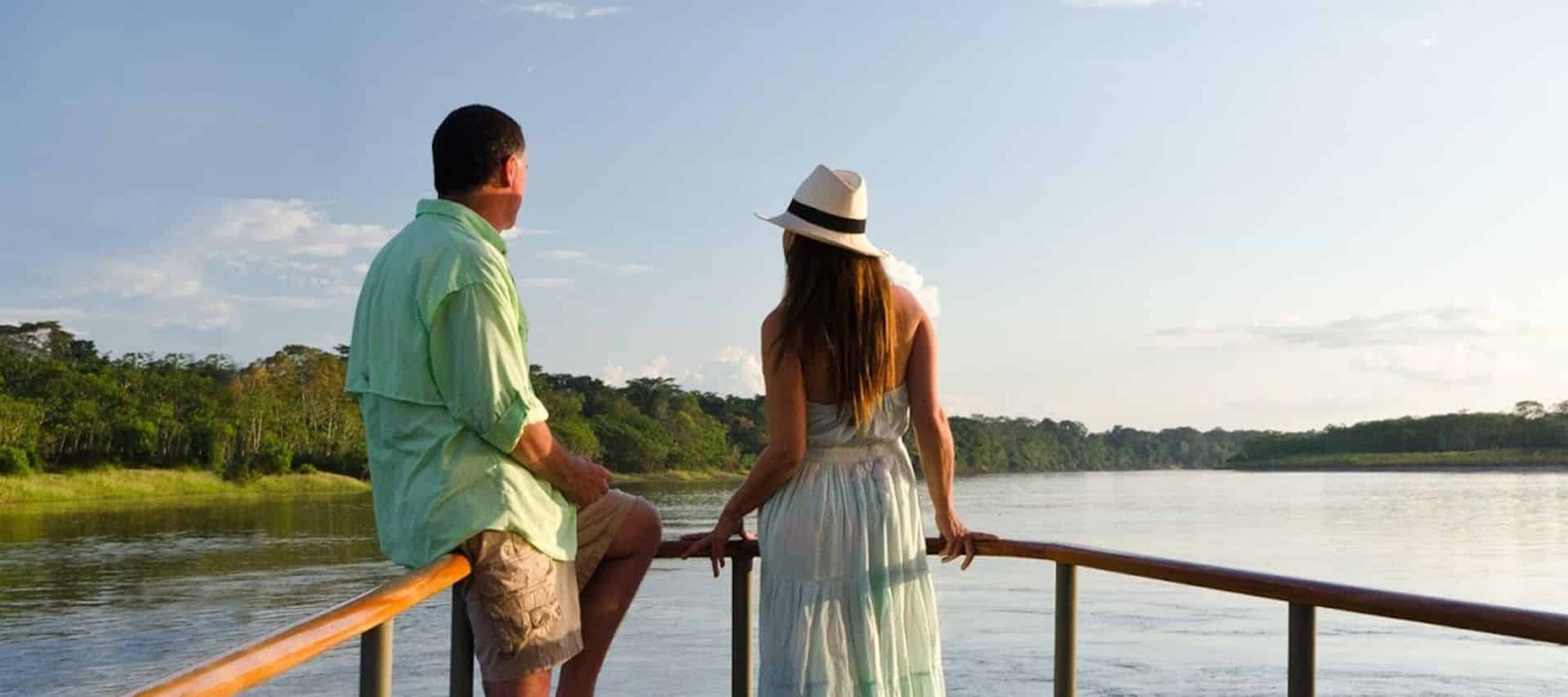 A man and a woman sit on the railing of a boat, looking out over the calm Amazon River in Peru, surrounded by lush greenery under a clear, sunny sky. The woman wears a hat and light dress; the man wears a light shirt and shorts—perfect for river cruises.
