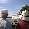Two people wearing hats sit in a boat on a river, facing away from the camera, enjoying an unforgettable trip as they watch a white riverboat pass under a blue sky with scattered clouds and trees lining the riverbank.