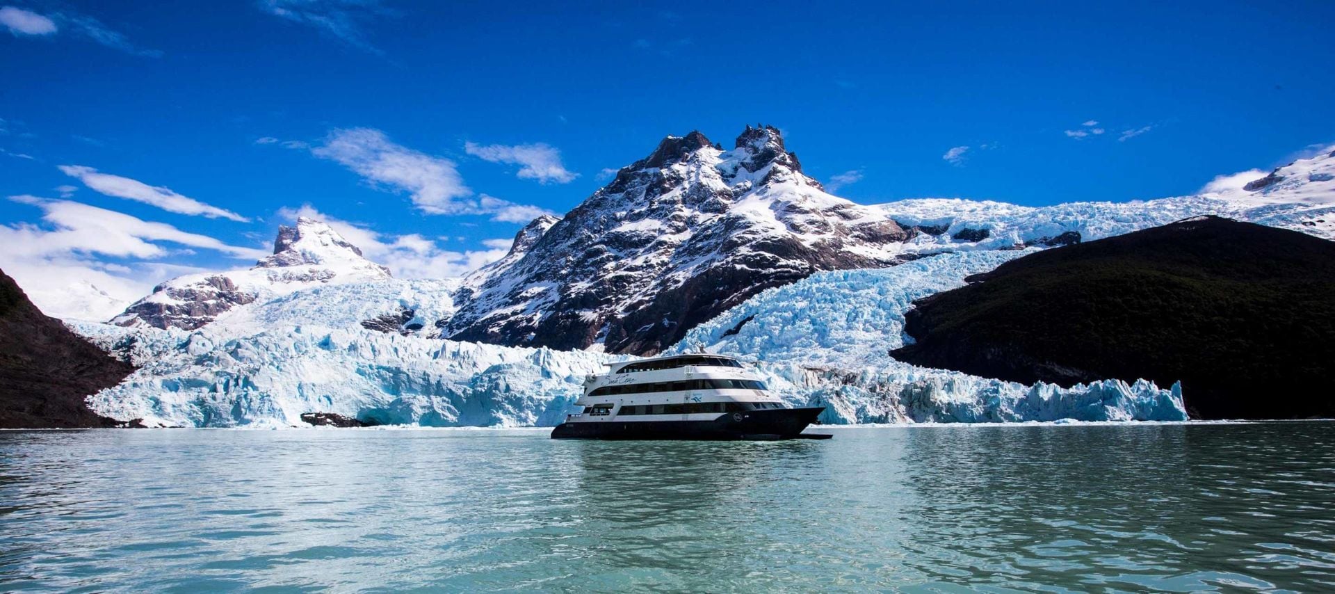 A large boat sails on a calm, icy blue lake with snowy mountains and a glacier in the background under a clear blue sky, offering an unforgettable experience typical of Chile cruises.