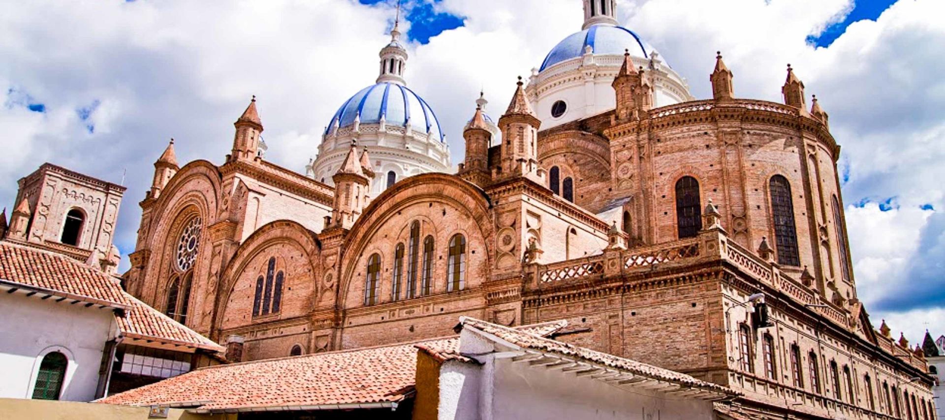 Exterior view of a large cathedral with three blue-and-white domes, arched windows, and detailed brickwork under a partly cloudy sky. A highlight of Cuenca Tours, the building combines Gothic and Renaissance architectural elements.