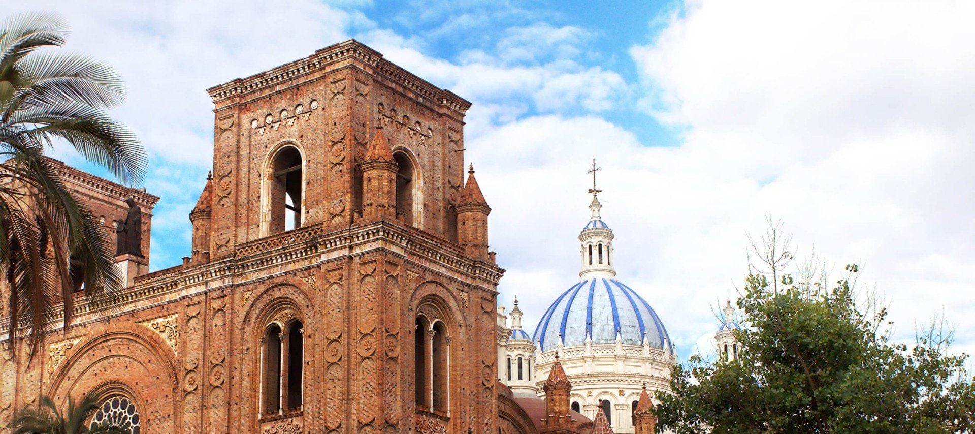 A historic red-brick cathedral with arched windows stands in front of a blue-domed church under a partly cloudy sky, surrounded by palm and leafy trees—a striking scene often admired on UNESCO tours in Ecuador.
