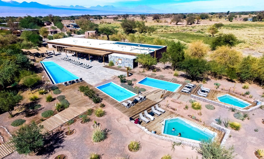 Aerial view of Cumbres Atacama resort and spa with multiple pools, lounge chairs, wooden decks, and desert landscaping, surrounded by trees and fields, with mountains visible in the distant background.