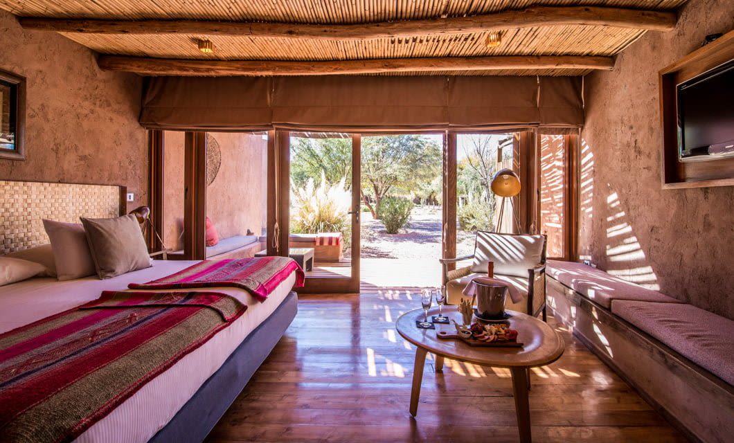 Cozy, sunlit bedroom at Cumbres Atacama with large glass doors opening to a garden. Features include a bed with a colorful blanket, cushioned bench, wooden ceiling, snacks and drinks on a small table, and views of trees outside.