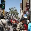 A busy city street scene with many people walking on the sidewalk. Shops known for their superb service line the street, and a green pedestrian traffic light is visible. The sky is clear and blue.