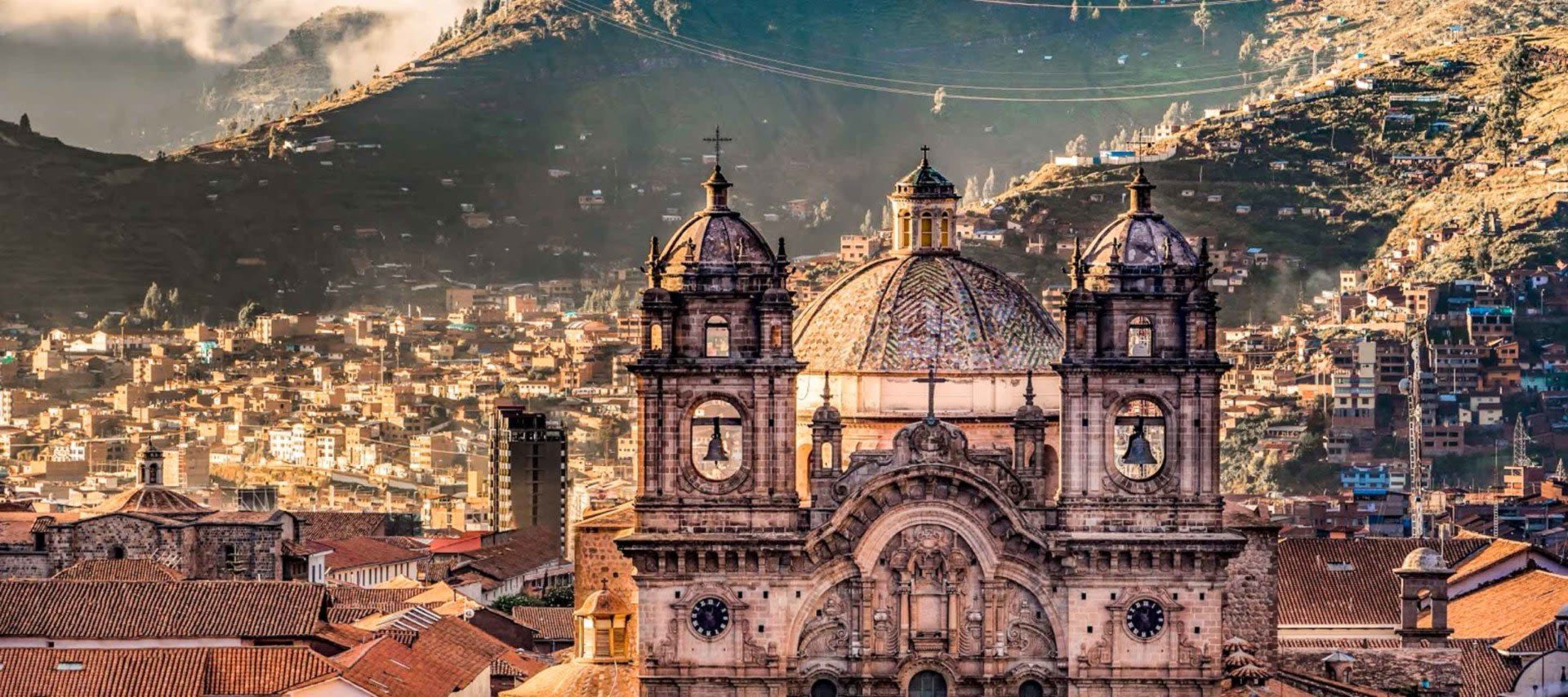 A historic stone cathedral with two towers stands among red-tiled rooftops, set against a densely built hillside and rugged mountains in the background under a cloudy sky.
