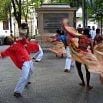 A group of dancers in colorful outfits perform energetically in an outdoor plaza, their blurred motion capturing a fantastic time. Trees and a large stone monument are visible in the background.
