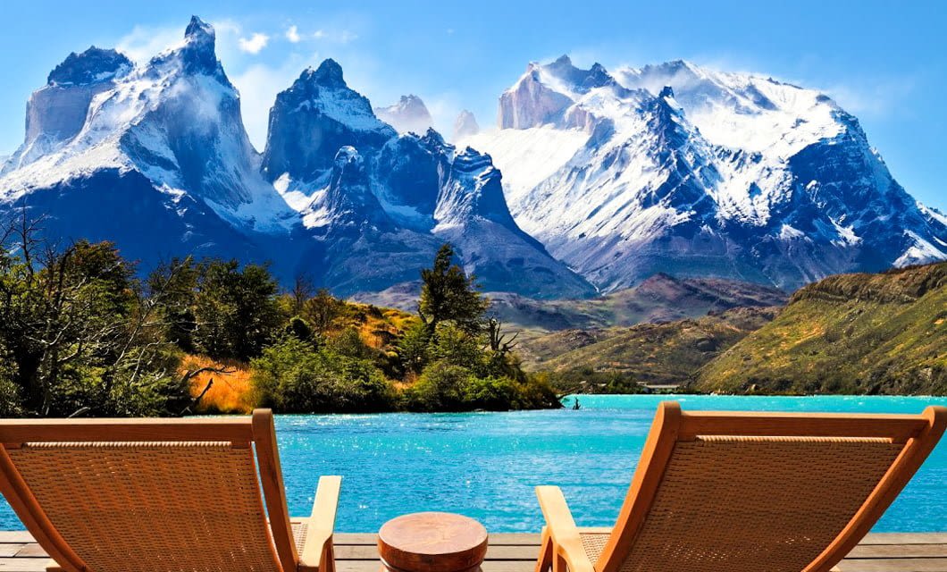 Two wooden lounge chairs face a bright turquoise lake with snow-capped mountains in the background under a clear blue sky, surrounded by greenery and rugged scenery near Hotel Las Torres.