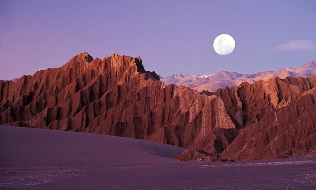 Jagged, reddish-brown mountains under a purple sky at dusk evoke the tranquil beauty of Awasi Atacama, with a full moon rising above distant peaks and a flat, shadowed plain in the foreground.