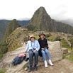 Two people sit on a stone wall at Machu Picchu, Peru, with the iconic mountain and ancient ruins in the background. Both wear hats and casual clothing, enjoying their perfect trip after carefully arranging trip details for this scenic, cloudy day.