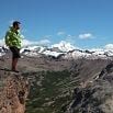 A person in a bright green jacket stands on the edge of a rocky cliff, having a terrific time as they overlook a vast landscape of mountains with snow-capped peaks under a clear blue sky.