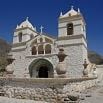 A historic white stone church with two bell towers stands under a clear blue sky, surrounded by mountains and rocky terrain—a perfect glimpse into South America for your next travel adventure.