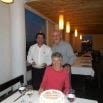 Three people pose together in a warmly lit Patagonia restaurant. A woman sits at a table with a cake in front of her, while two men stand behind her. Hanging lights and set tables evoke the cozy ambiance of Skorpios III gatherings.