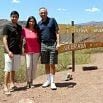 Three people stand smiling in front of a wooden sign on a dirt path, with grassy hills and mountains behind them under a blue sky—a perfect snapshot of a good trip.