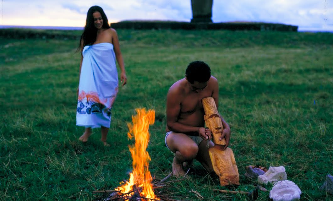 A man kneels on grass near a small campfire, carving a wooden statue, while a woman in a white wrap dress stands nearby. In the background, the iconic Moai statues of Rapa Nui rise over Easter Island, creating an atmosphere unique to Hare Noi.