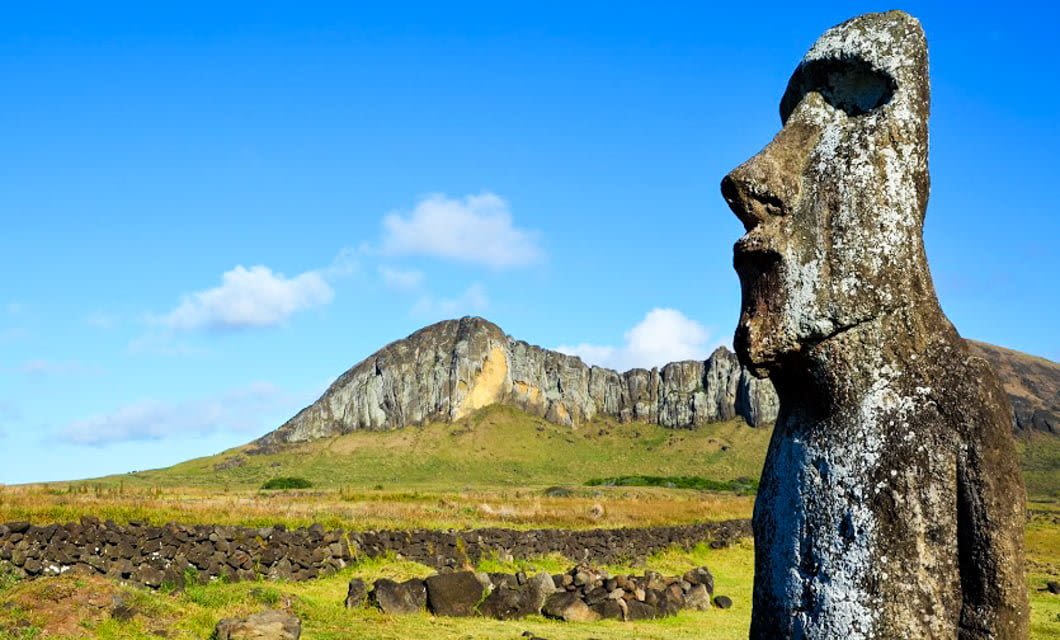 A large stone Moai statue stands in the foreground on Easter Island, or Rapa Nui, with grassy fields, stone walls, and a rocky hill under a bright blue sky in the background—perfect for an Explora adventure.