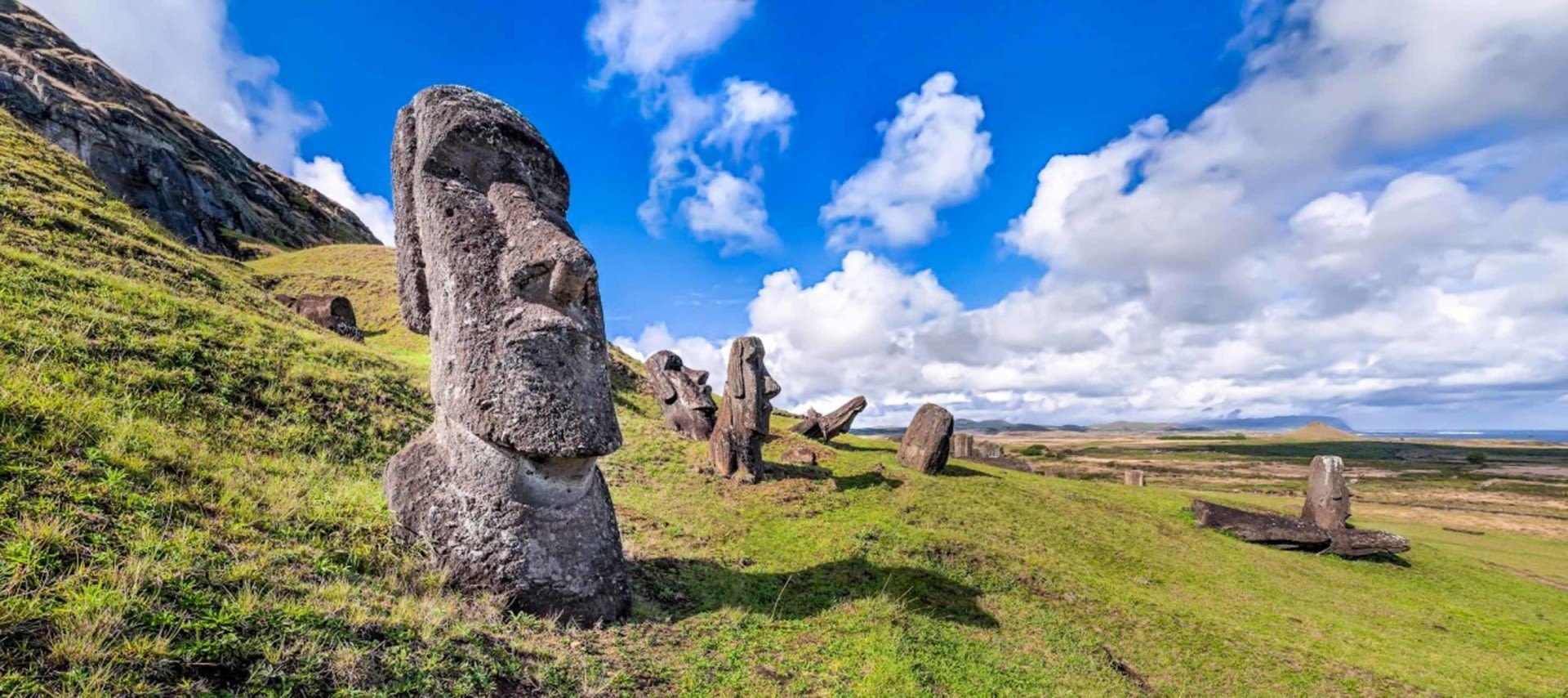 Large stone Moai statues stand on a grassy hillside under a bright blue sky on Easter Island, surrounded by rolling landscape and distant mountains—an iconic sight often explored on Easter Island Tours.