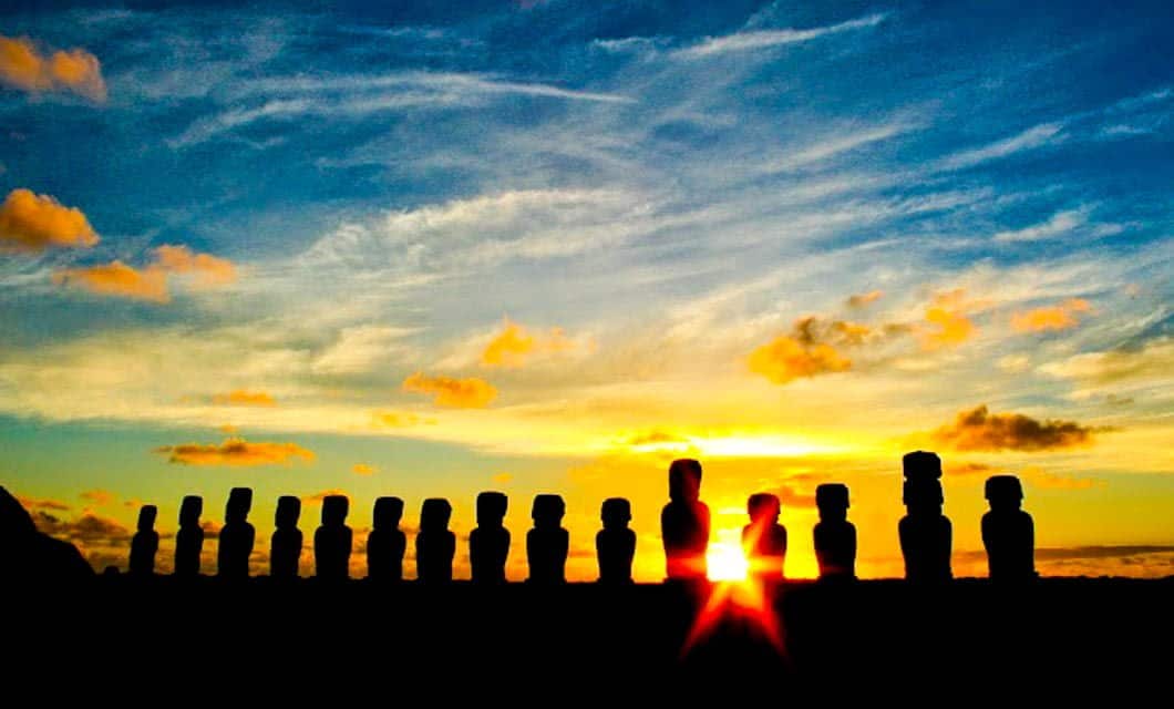 Silhouetted moai statues on Easter Island stand in a row against a vibrant sunset sky, with the sun shining between two of the statues and clouds scattered across the colorful horizon near Hangaroa Eco Village, an eco-friendly hotel.