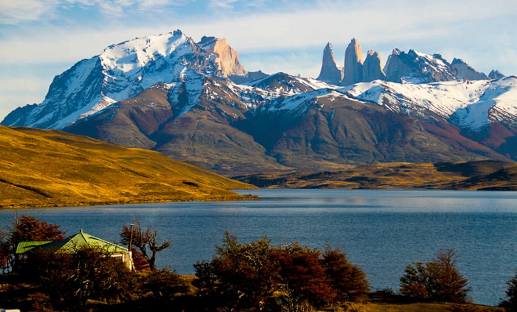 Snow-capped mountains rise above a serene blue lake, with golden grassy hills and scattered trees in the foreground. An eco-friendly Patagonia Lodge with a green roof sits near the shore under a partly cloudy sky.