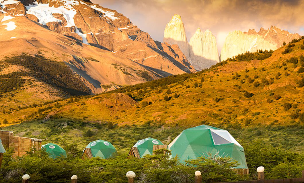 Green geodesic dome tents at this Eco-Camp lodge are set on a hillside with scattered bushes, below Patagonia’s rugged mountains aglow with golden sunlight, beneath a partly cloudy sky.