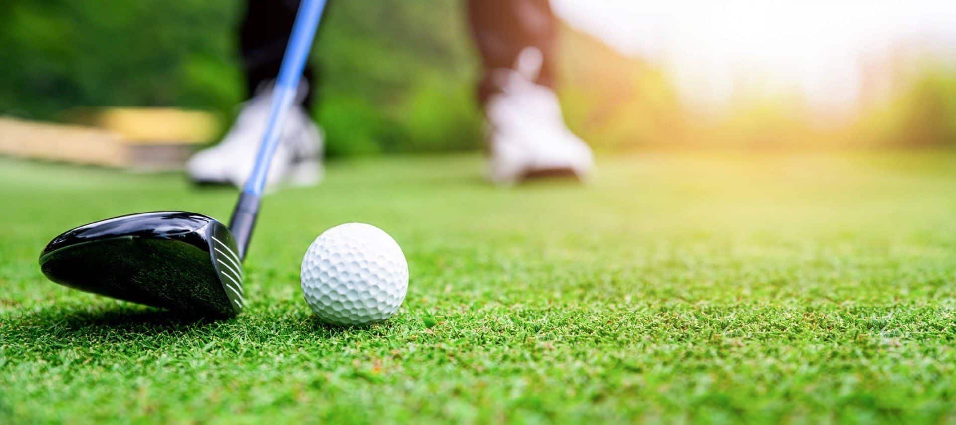 A close-up of a golf club about to hit a golf ball on a green course in Ecuador, with a person’s legs and shoes in the background and sunlight illuminating the scene.