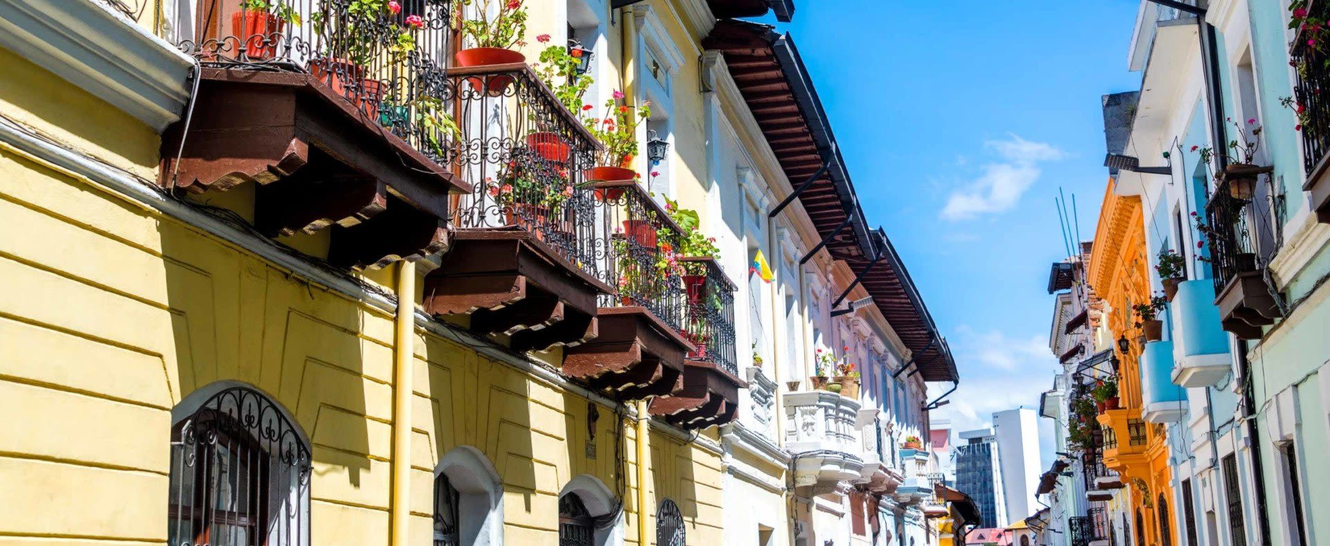 Colorful colonial-style buildings with ornate iron balconies and potted plants line a narrow street under a bright blue sky, capturing the charm of Ecuador travel and offering a glimpse into the vibrant culture visitors experience after meeting Ecuador visa requirements.