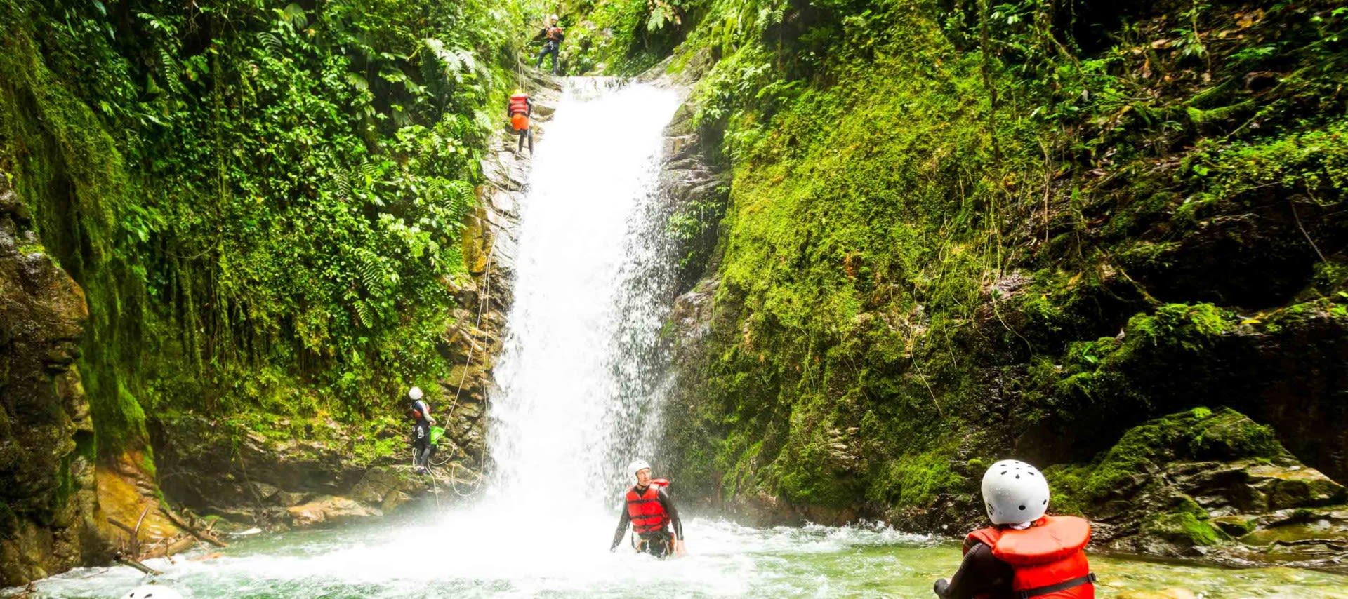 People wearing helmets and red life vests stand and climb near a lush, green waterfall in the jungle—an exciting scene from South America Tours, where adventure awaits amid dense vegetation and rocky waterfalls.
