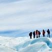 A happy group of people wearing winter clothing stand on a snowy and icy landscape under a cloudy sky, exploring the glacier with their guide during a thrilling tour.