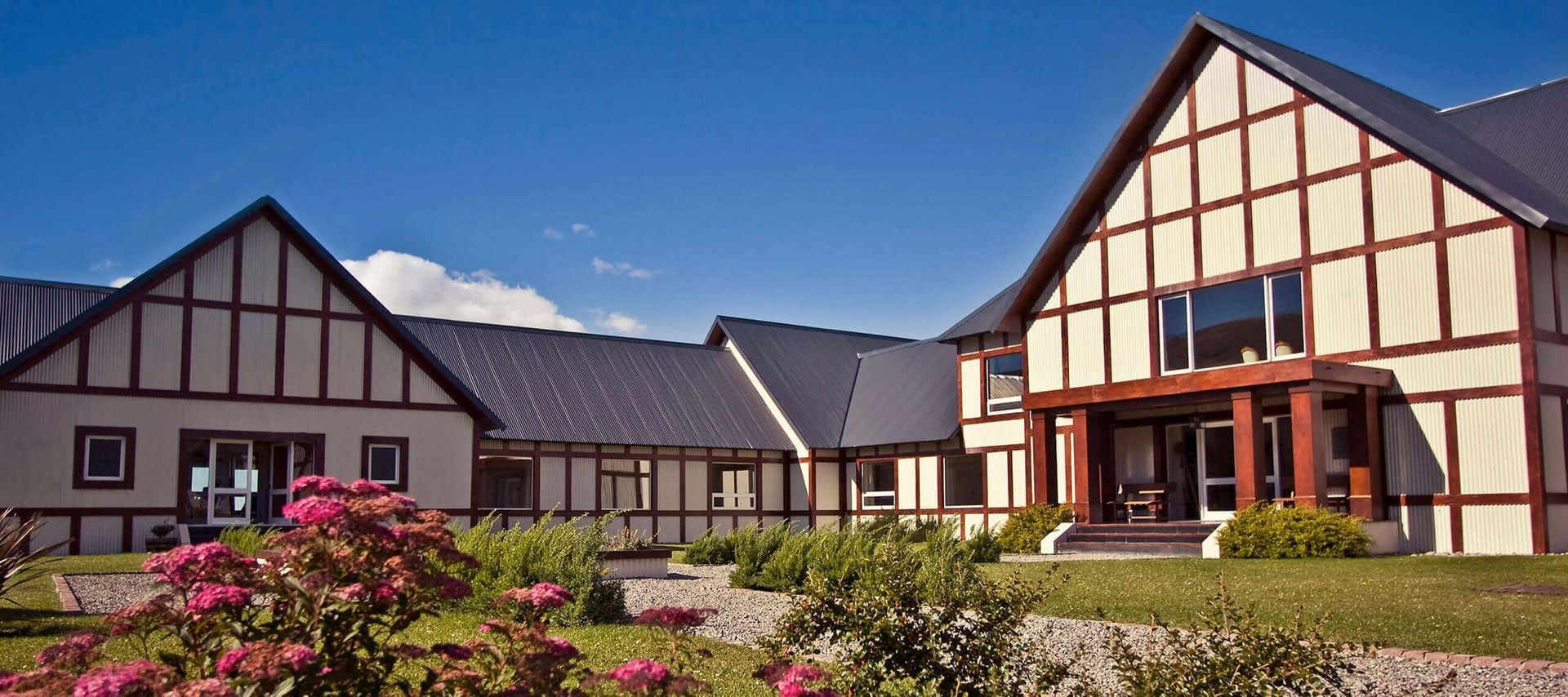 A modern building with Tudor-style architecture, steep black metal roofs, and white walls with dark wooden beams, reminiscent of Argentina lodges, sits on a front lawn with flowering plants under a bright blue sky.