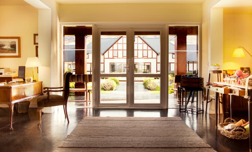 A bright, elegant room at Eolo Patagonia features wooden furniture, a large rug, and double glass doors opening to a garden courtyard with views of a traditional-style building outside.