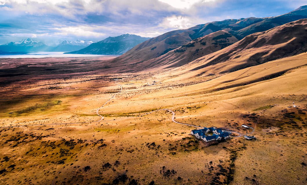Aerial view of Eolo Patagonia Lodge, a remote house surrounded by rolling golden hills, winding paths, and sparse vegetation, with mountains and a lake in the distance under a cloudy sky.