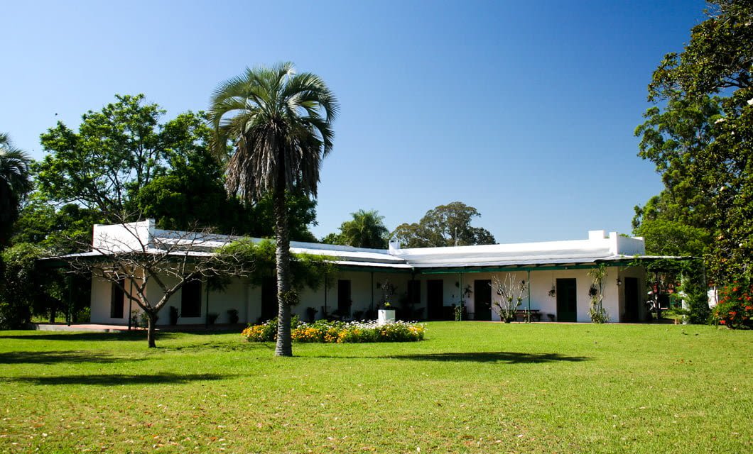 Estancia Buena Vista is a white, single-story house with a flat roof, surrounded by lush green lawn, palm trees, and flowering plants under a clear blue sky.