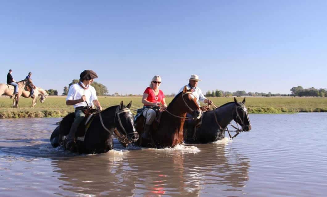 Three people riding horses through a shallow river under a clear blue sky, with grassy fields and two other riders in the background—a perfect example of adventurous things to do in Uruguay.