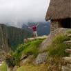 A person stands with arms raised on a grassy ledge near a stone structure with a thatched roof, overlooking misty mountains and valleys—capturing memorable moments from an amazing trip.