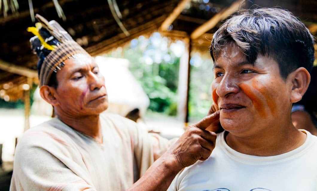 An Indigenous man in traditional attire paints orange lines on another man's face under a thatched-roof shelter at Corto Maltes Lodge, surrounded by a lush, vibrant outdoor setting.