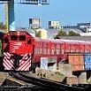 A bright red passenger train travels along train tracks in Buenos Aires, passing buildings and industrial structures under a clear blue sky.