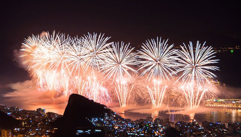 Fireworks burst in the night sky above a South American city and waterfront, illuminating the buildings below with bright white and gold light—a stunning New Year's Eve celebration in one of the best cities. Smoke from the fireworks drifts over the water and city.
