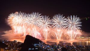 Fireworks burst in the night sky above a South American city and waterfront, illuminating the buildings below with bright white and gold light—a stunning New Year's Eve celebration in one of the best cities. Smoke from the fireworks drifts over the water and city.